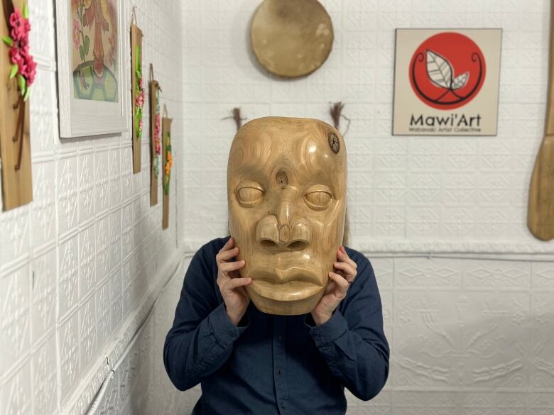 A man holds a large, wooden mask in front of his face.