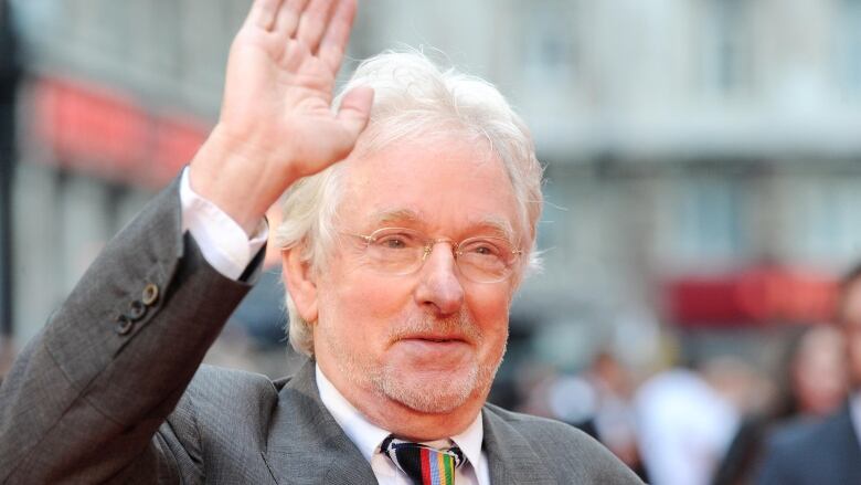 A white-haired man wearing glasses waves.