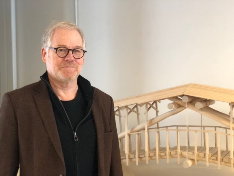 Man in brown blazer wearing glasses stands beside model of a wooden bridge.