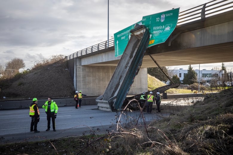 A commercial truck trailer is tilted up against an overpass above a highway. First responders are gathered on the ground.