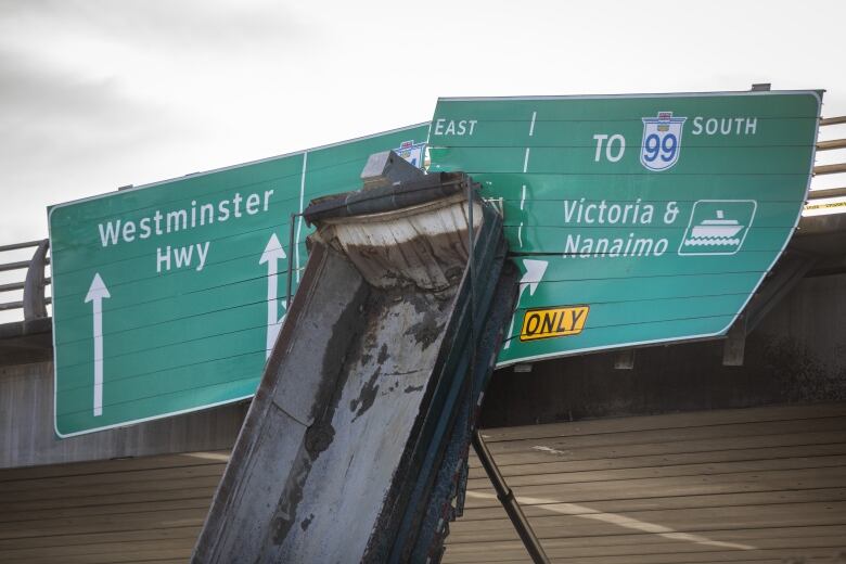 A commercial truck trailer leans on highway signs hanging from an overpass.