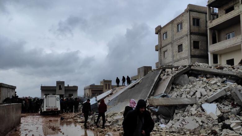 Local people in Turkey are shown standing and walking in front of a fallen building. 