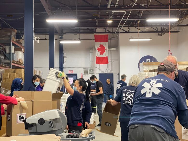 Many people are shown lifting buckets and moving boxes. Most are wearing blue GlobalMedic t-shirts and Canada flag hangs in the background.