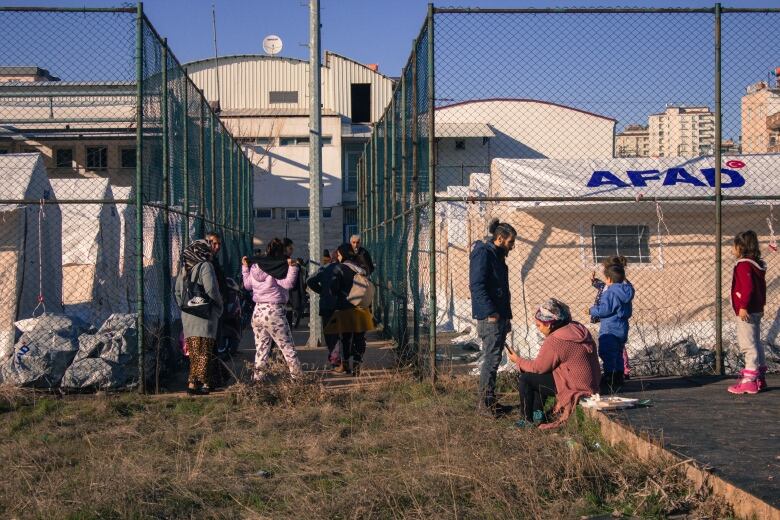 A handful of Turkish people are shown standing outside a group of tents enclosed behind chain link fences.