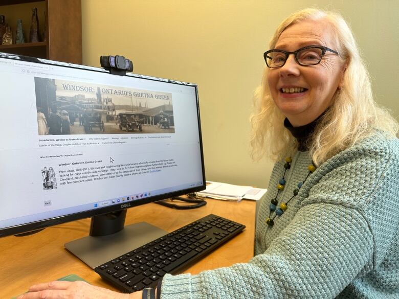 A woman sits beside a computer screen with a website up.