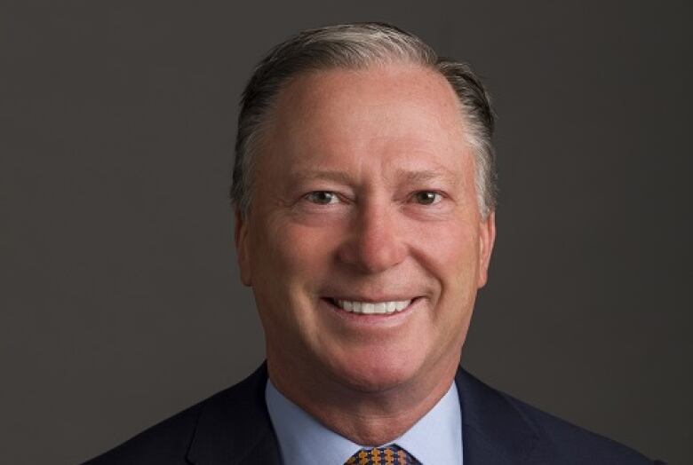 A man poses for a photograph wearing a dark suit and a tie. He is smiling.