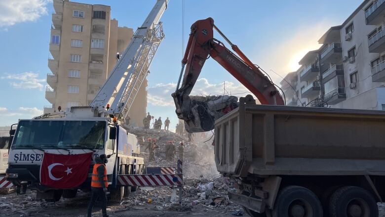 Two construction vehicles in operation dig through rubble as workers watch from a distance.