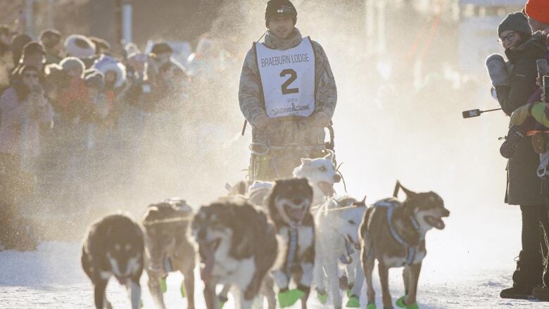 A musher is seen on a sled with eight dogs, people are cheering around him. 