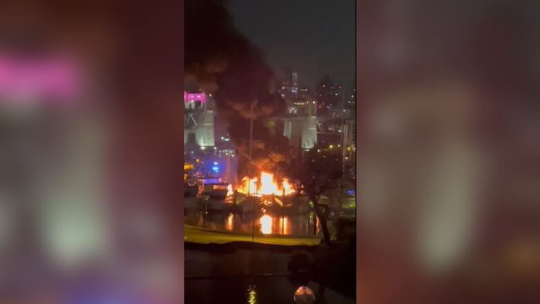 A large fire is seen on the water in a city, with emergency lights flashing and a large plume of smoke visible.