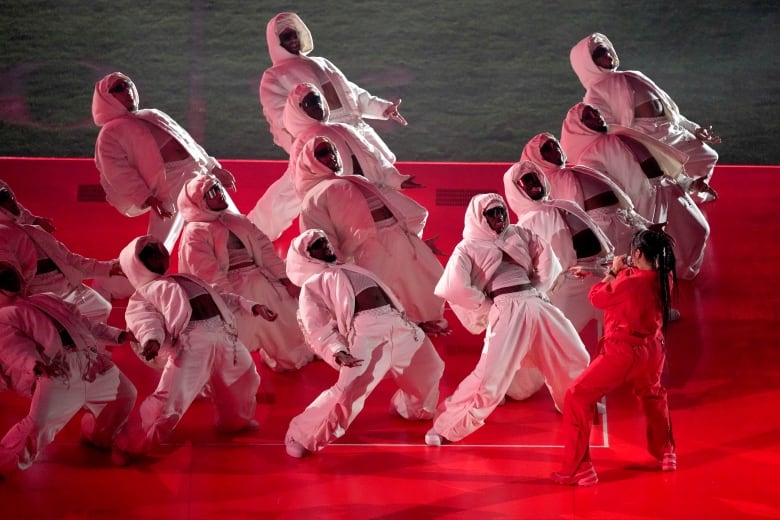 A group of dancers wearing white stand opposite a woman wearing red.