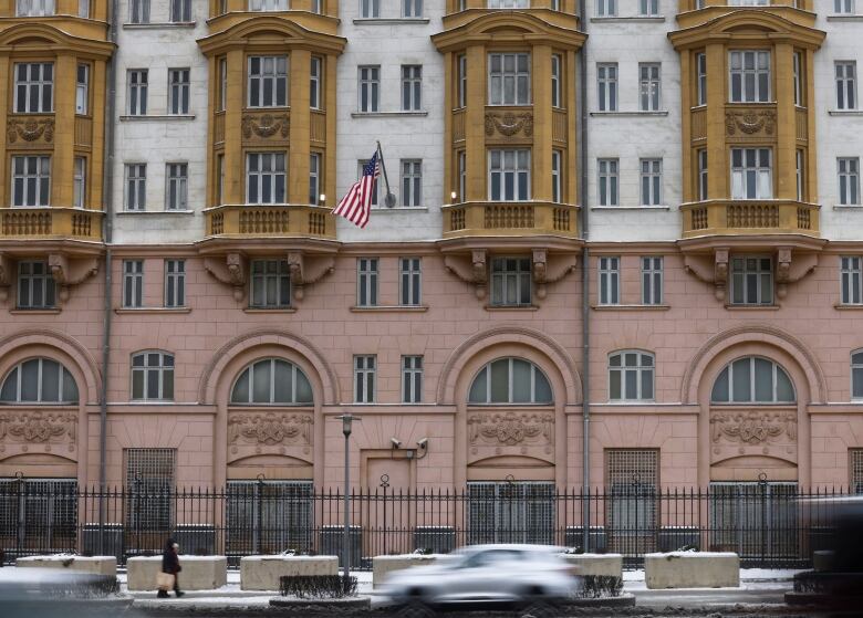 A multi-storey building behind a black fence and with many windows is seen from a distance across a city street. An American flag hangs diagonally from the fourth storey.