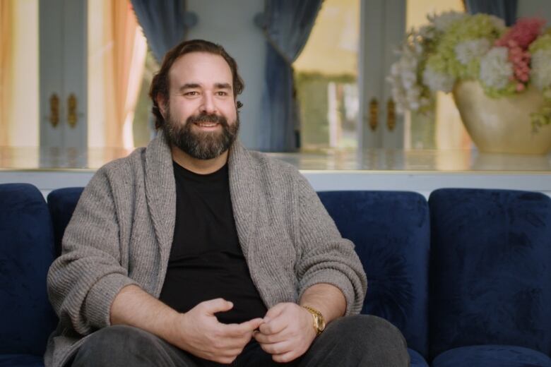 Bearded man wearing a gray cardigan speaks on camera while sitting on a blue sofa.