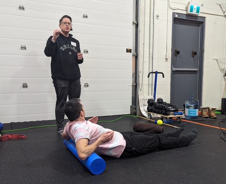 A person lies on the ground using a roller behind their back, while another trainer stands above speaking to a crowd.