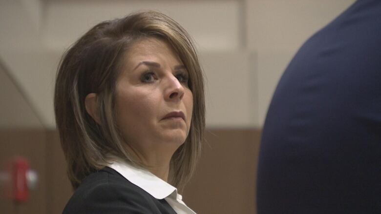 A woman wearing a black suit and white shirt glances towards the camera, looking above it. 