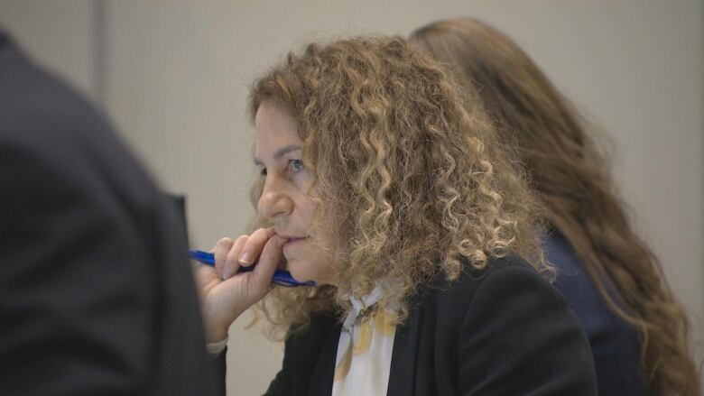 A woman in formal business attire bites her nails while holding a pen and looking forwards. 