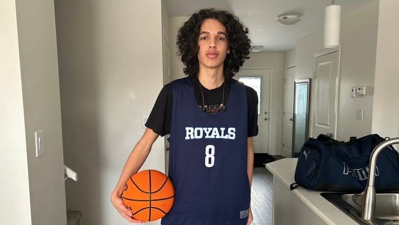 A tall teenage boy with long, poufy hair, wearing a blue number 8 Royals basketball uniform over a black t-shirt is pictured while holding a basketball in the kitchen of a home.