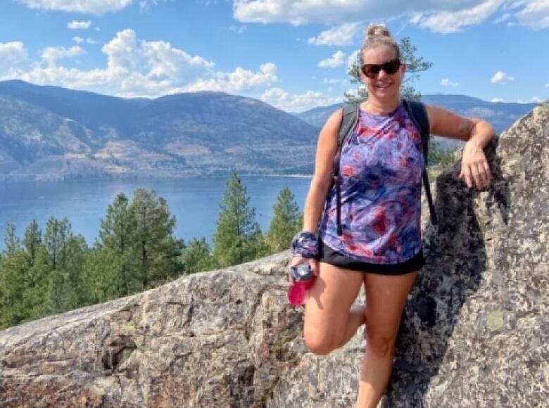 A woman in shorts and a blue T-shirt leans on a rock as she poses for the camera. In the background, there are mountains and a lake.