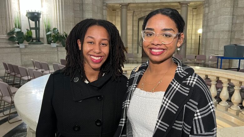 Two Black teenagers smile for the camera in a marble room. The girl on the left is wearing a black jacket, while the girl on the right is wearing a black and white plaid jacket, a white top, and clear-framed glasses.