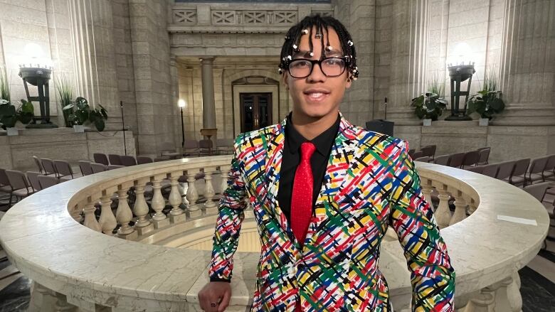 A Black teen with his hair in braids smiles at the camera. He's wearing black-framed glasses, and a colourful suit jacket with a black button-down and red tie.