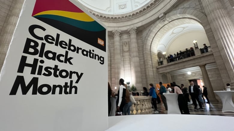 A number of people stand around tables in the background in front of a sign marked with the Manitoba NDP logo that says 