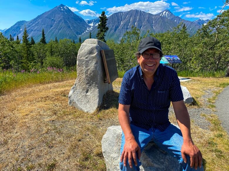 A smiling man sits on a rock on a sunny day, with mountains visible in the background.