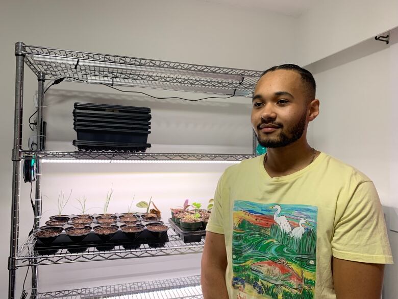 A man stands beside several small plants in multiple containers on small stainless steel shelves.