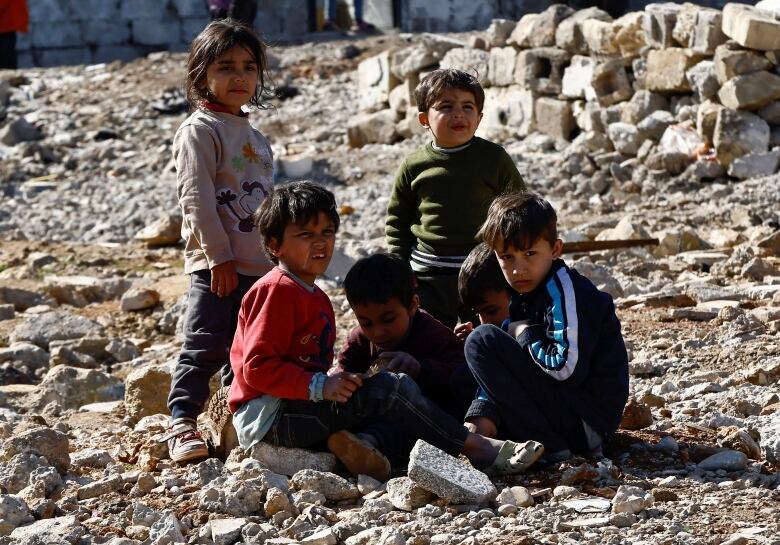 Six children are seen either sitting or standing outside on ground covered with rocks and stones.