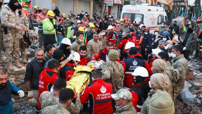 Several rescue crew members dressed in red are shown, some carrying a stretcher in the direction of an ambulances amid rubble and a crowded urban setting.