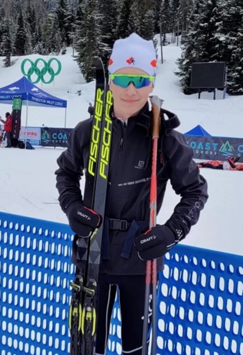 A teenaged boy holds his cross-country skis and poles in front of blue netting around a snowy cross-country ski track.