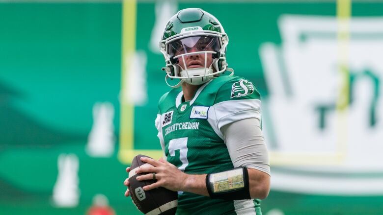 A quarterback in a green jersey holds a football in his hands as he steps back to pass.