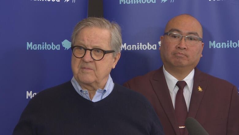 A man in a dark sweater over a collared shirt stands next to another man in a maroon-coloured blazer and tie at a news conference.