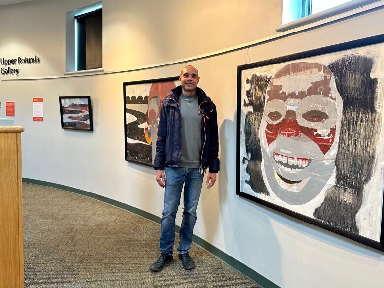 A male Black artist stands next to a smiling, colourful, tribal inspired artwork on display at the Upper Rotunda Gallery in Richmond, B.C.