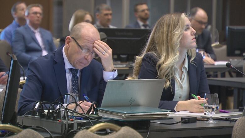 A woman and man seated at a table in a hearing room. The man is writing on a legal tablet.