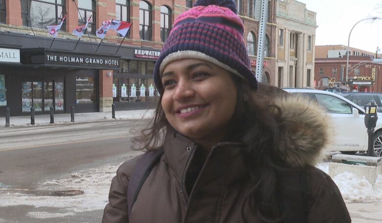 Woman in a heavy winter coat and woollen hat smiles while standing on a downtown Charlottetown sidewalk. 