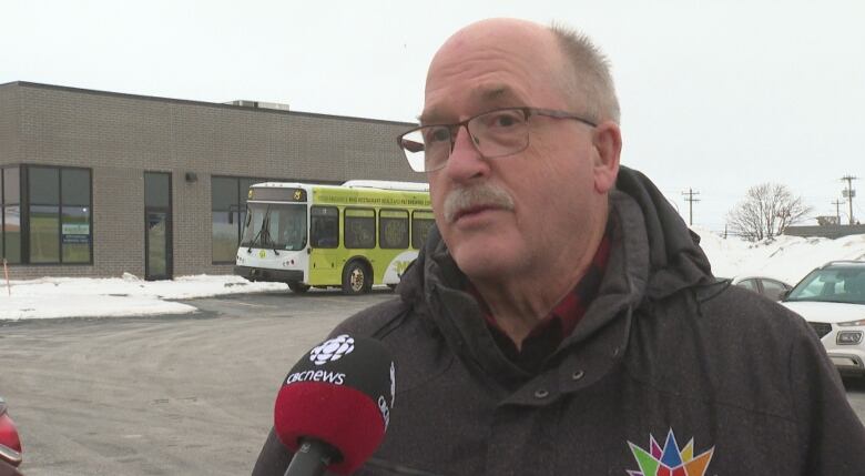 Man wearing glasses and a black jacket talks into a microphone in a parking lot with a T3 bus parked behind him. 