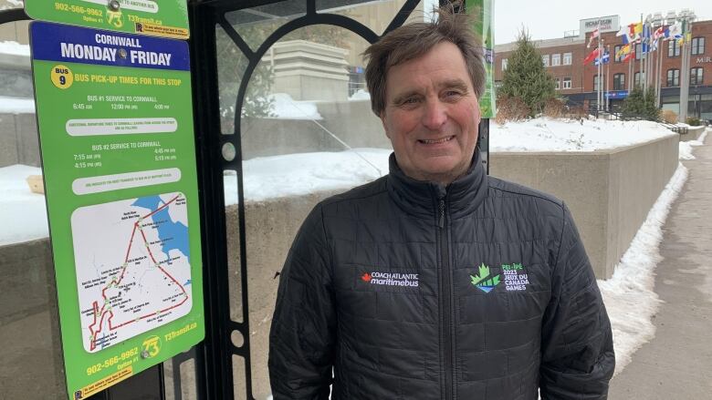 Smiling man in black jacket stands in front of a bus stop with snow in the background. 