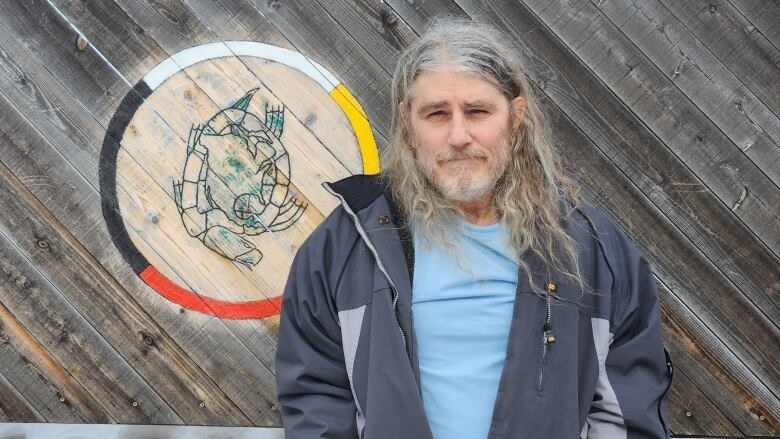 A man with long grey hair wearing a blue shirt and grey jacket stands with his hands in his pockets in front of a wooden wall.