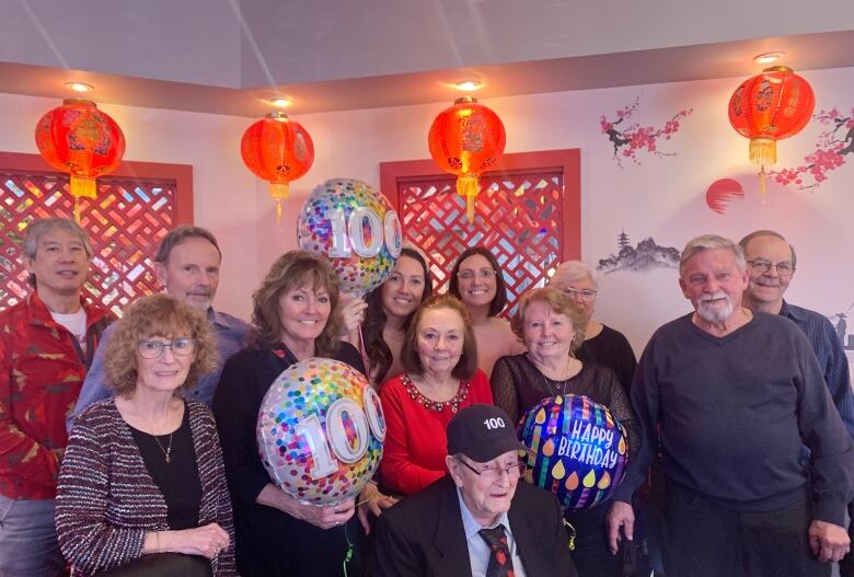 A group of people, some holding happy birthday balloons, surround an elderly man in a hat inside a Chinese restaurant with lanterns hanging from the ceiling 