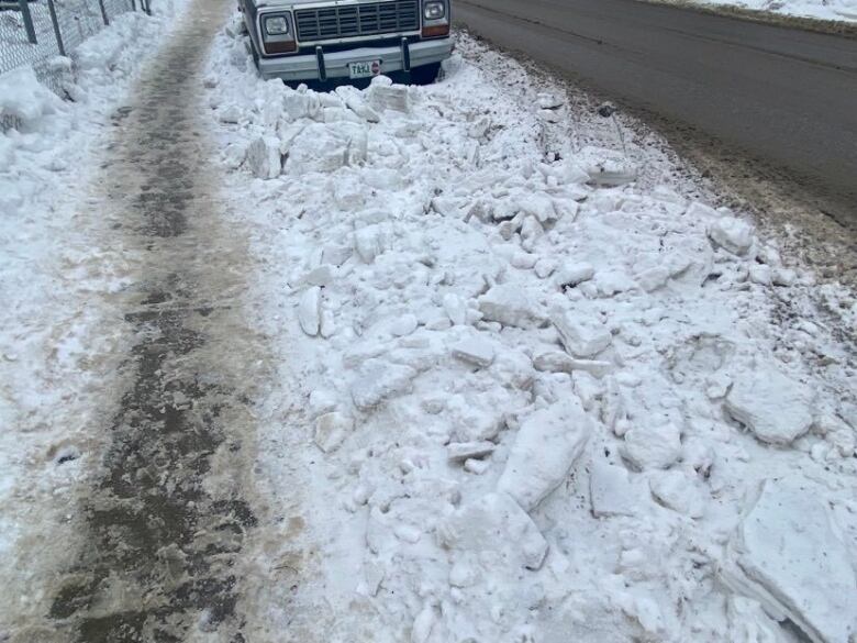 A thin strip of clear sidewalk with snow on the side and piled high in the road in front of a truck.