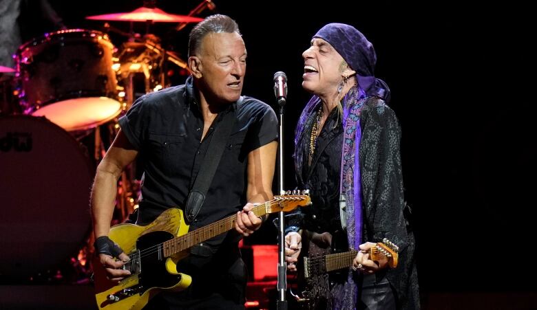 Two men holding guitars sing into a single microphone on a stage.