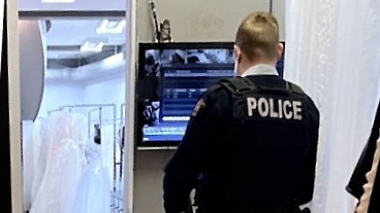 A man with Police emblazoned on his dark uniform and a yellow stripe running down the side of his pants looks at a computer screen in a bridal shop with wedding dresses in the background.