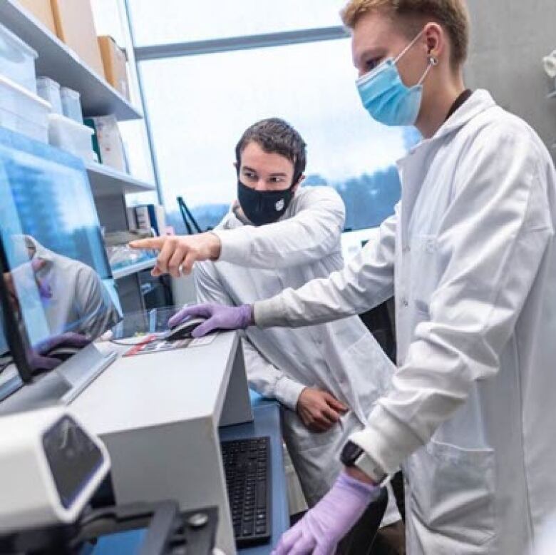 Two people in white coats wearing medical masks point at the screen of a computer monitor as they discuss their research.