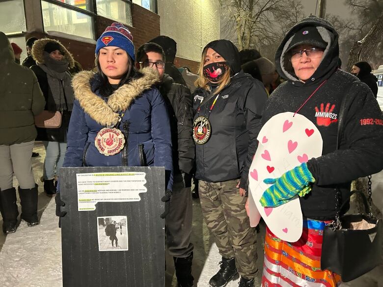 Four people pose for a photograph on a snow-covered sidewalk, bundled up in winter clothing holding signs bearing the names of missing and murdered women.