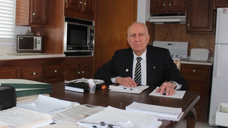 Older man in a suit sitting at his kitchen table, which is covered with stacks of paper and files