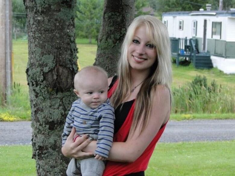 Young smiling woman with blonde hair holding a baby and posing for a photo outside in front of a tree