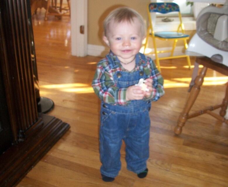 Blonde smiling toddler wearing a plaid shirt and overalls standing in the middle of a dining room looking at the camera