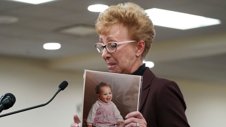 An emotional older woman holds up a photo of a baby girl while standing at a lectern.