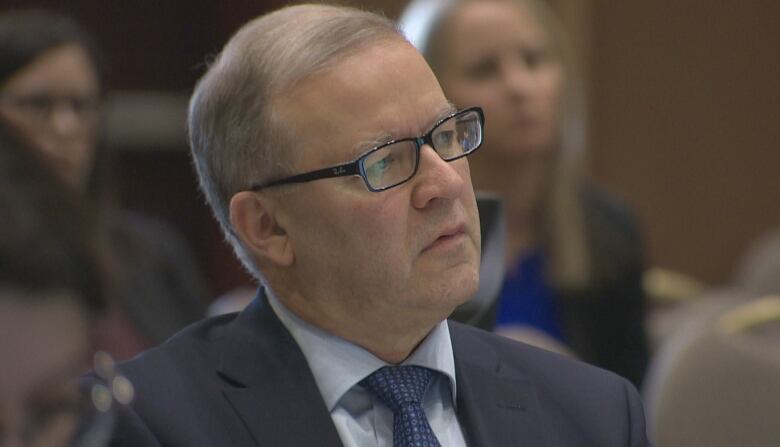 A man wearing glasses and a suit and tie. There are people sitting behind him.