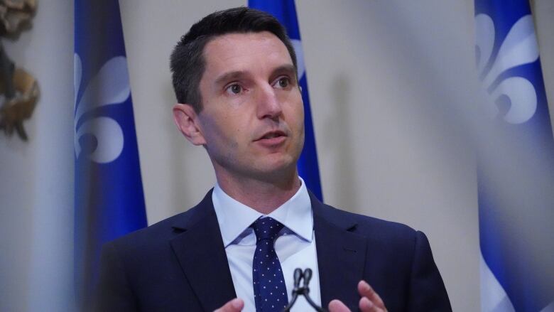 A man in a tie speaks in front of Quebec flags. 