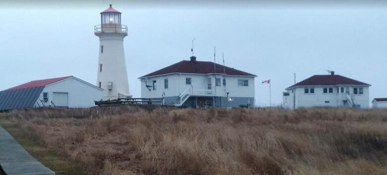 A lighthouse and several outbuildings with a wooden boardwalk leading over long grasslands leading up to the buildings.